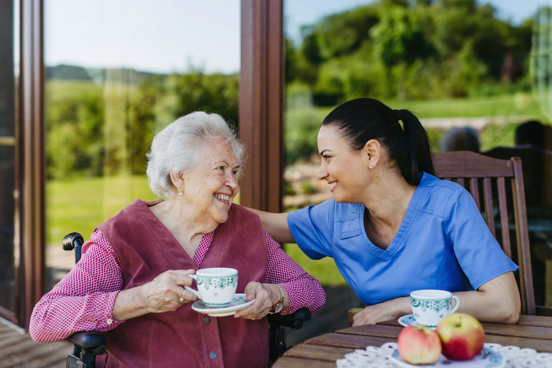 Female caregiver spending time with elderly woman, drinking coffee and talking. Nurse and senior woman in wheelchair enjoying a warm day outdoors.